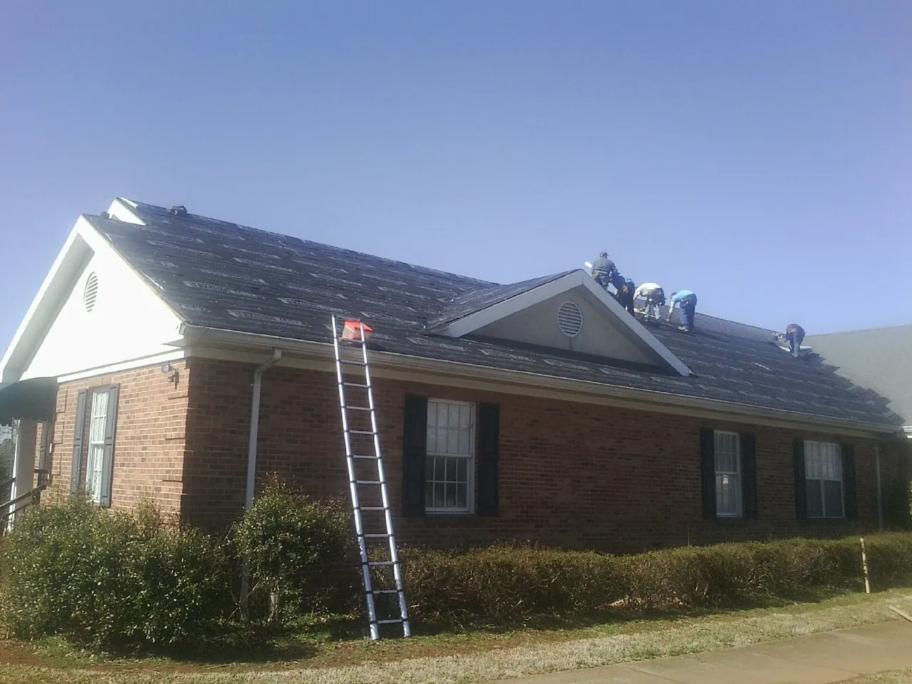 Crew tearing shingles off a brick ranch roof with a ladder set — Emergency Roof Repair work by our crew in Hurt