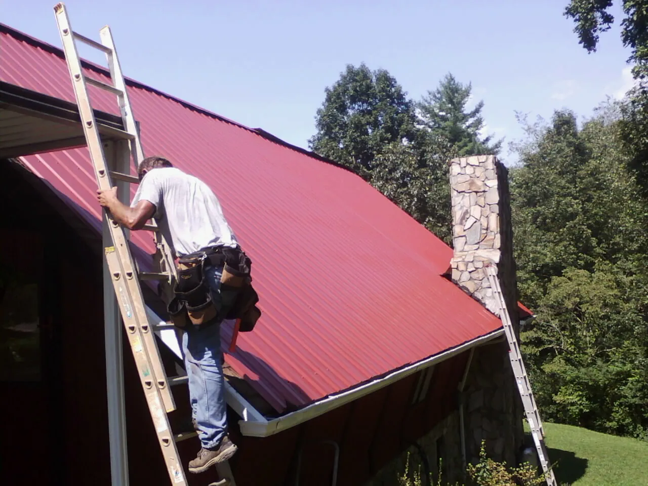 Roofer on a ladder beside a red metal roof and stone chimney — Soffit & Fascia Repair by Roofing Brothers in Hurt